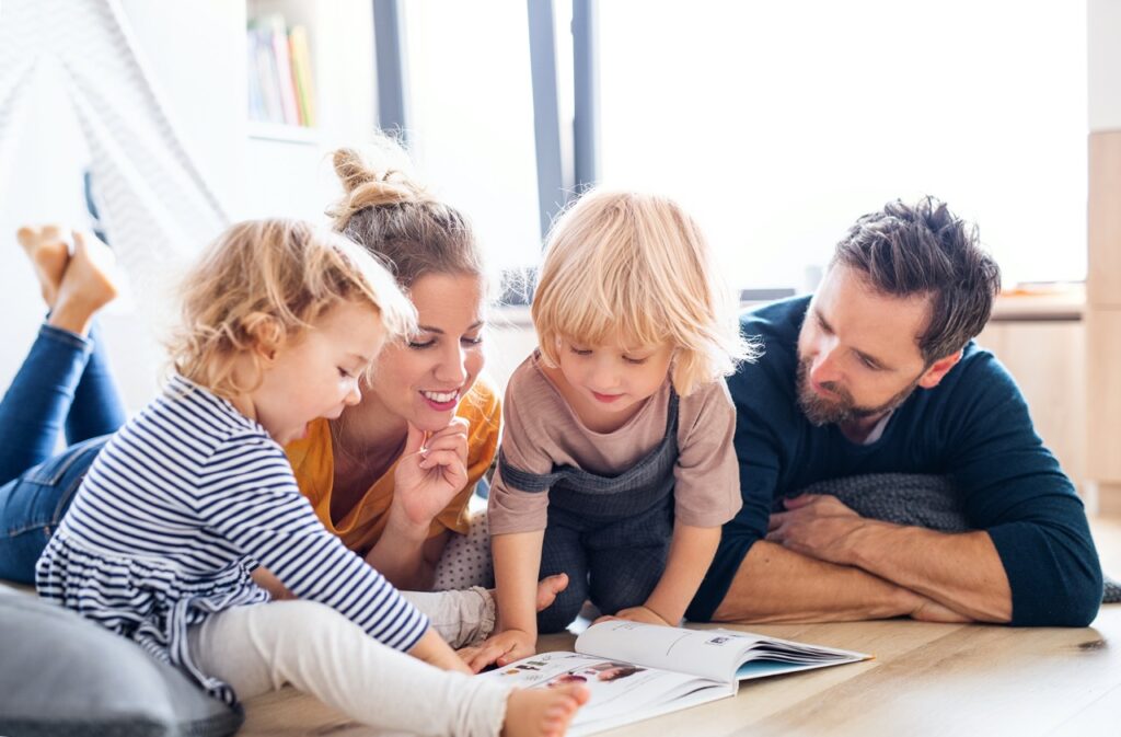 Front view of young family with two small children indoors in bedroom reading a book.